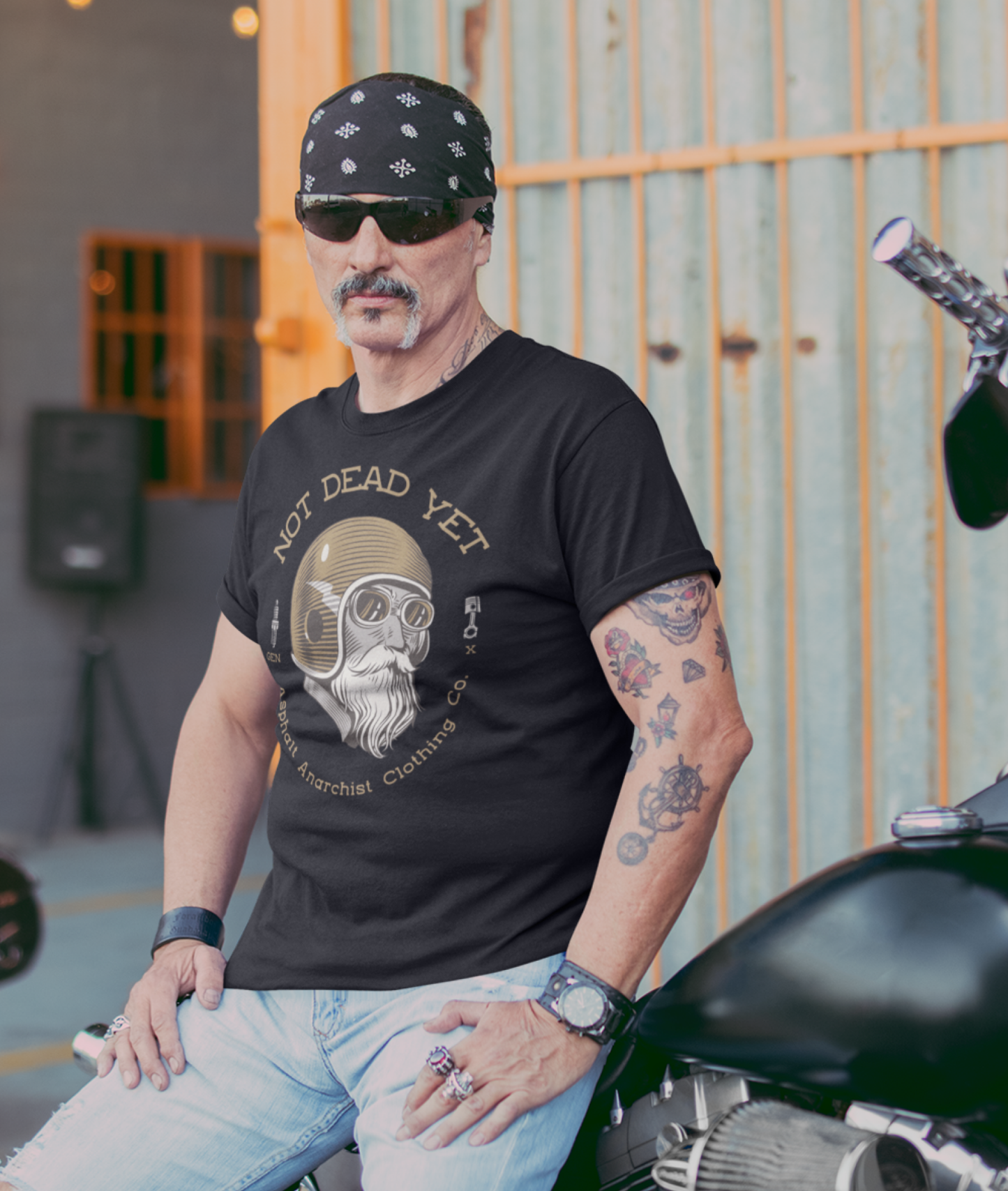 A crusty older biker leans on his hog. He is wearing a black t-shirt featuring bold ‘Not Dead Yet’ graphic with vintage lowbrow skull art and distressed lettering in a rebellious, hot rod style. Asphalt Anarchist Clothing Co. OLD SKOOL HOT ROD APPAREL Custom fueled, never factory filled. Image is AI-generated.
