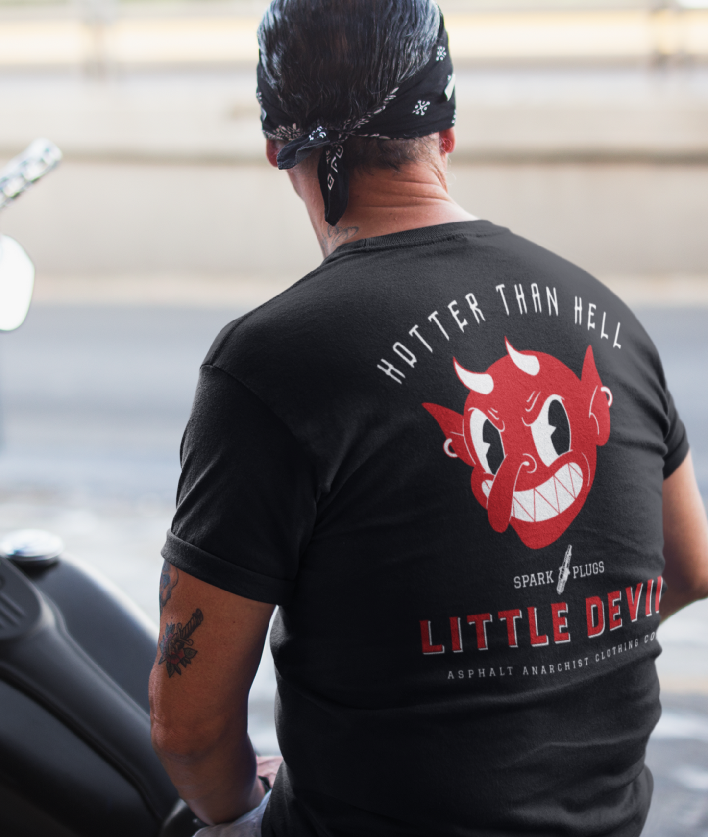 
                  
                    Little Devil Tee from Asphalt Anarchist Clothing Co. Black soft-style tee worn by an old-school biker posted on a Harley chopper. Retro devil head graphic and the slogan “Hotter Than Hell” blaze across the chest, echoing the gritty spirit of roadside garages and outlaw chrome. AI Generated image.
                  
                